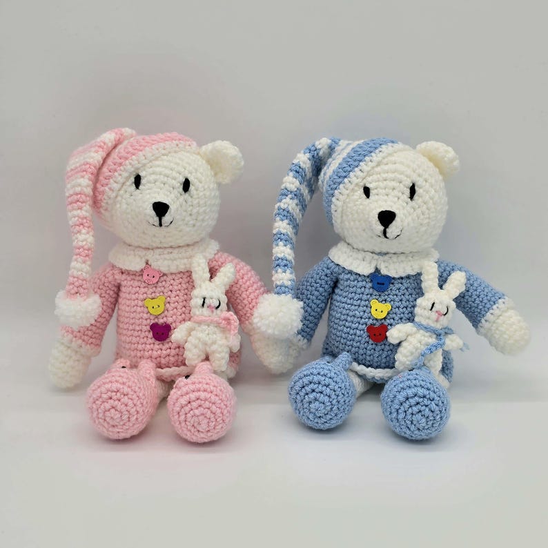 May include: Two crocheted teddy bears, one in pink and white and one in blue and white, each wearing a matching striped nightcap and pajamas with three colorful buttons. Each bear holds a small white crocheted rabbit toy on its chest.