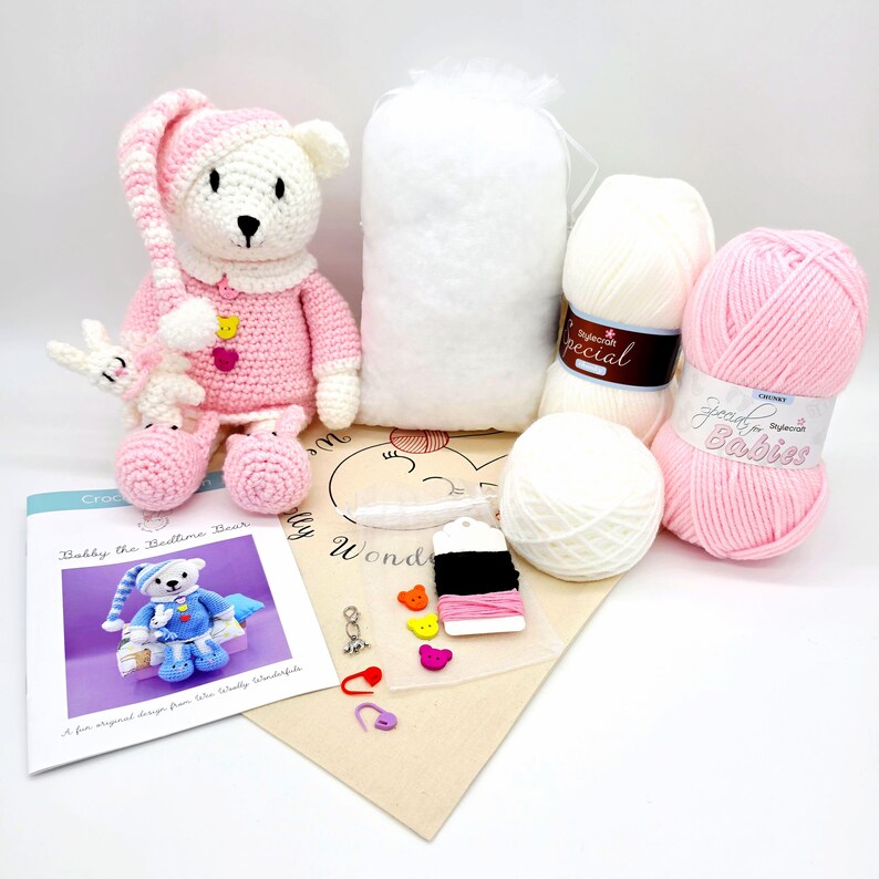 May include: A crochet kit featuring a finished white and pink bear doll, a bag of white stuffing, two skeins of white and pink yarn, a printed instruction booklet titled Bobby the Bedtime Bear, and a small packet of buttons, stitch markers, and thread.