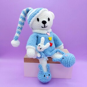 May include: A white crocheted teddy bear wearing a blue and white striped nightcap with a pom-pom, a blue sweater with three small colorful buttons, and blue bunny-shaped slippers. A small white crocheted rabbit is attached to the sweater front.