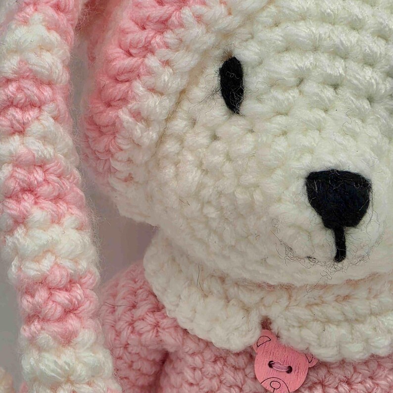 May include: A crocheted stuffed animal featuring a white head with a black nose and black eyes. It wears a white crocheted collar adorned with a light pink wooden button shaped like a bear face. The body and long ears are made of pink and white yarn.