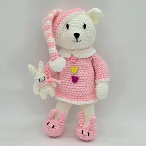 May include: A white crocheted bear toy wearing a pink and white striped nightcap, a pink dress with three bear-shaped buttons in pink, yellow, and purple, and pink bunny-shaped slippers. The bear holds a small white crocheted rabbit toy.