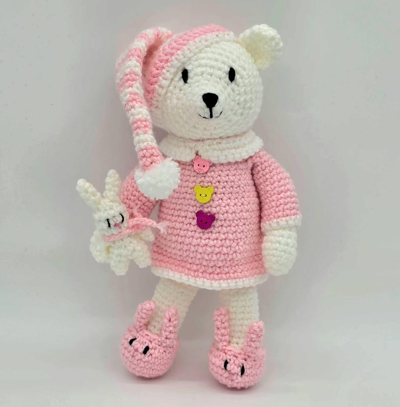May include: A white crocheted bear toy wearing a pink and white striped nightcap, a pink dress with three bear-shaped buttons in pink, yellow, and purple, and pink bunny-shaped slippers. The bear holds a small white crocheted rabbit toy.