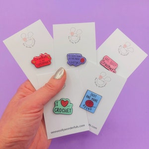 May include: Five enamel pins with different crochet-themed designs. The pins are on white cards with a bunny logo. The pins feature sayings like "Crochet is my superpower", "No such thing as too much yarn", "Eat, Sleep, Crochet", "I love crochet", and "Just one more round".