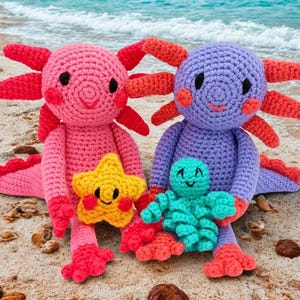 Puede incluir: Dos peluches de ajolote de ganchillo, uno rosa y otro morado, están en una playa de arena. El ajolote rosa sostiene una estrella amarilla. Un pequeño peluche de pulpo turquesa está frente al ajolote morado. Los ajolotes tienen branquias y pies rojos.