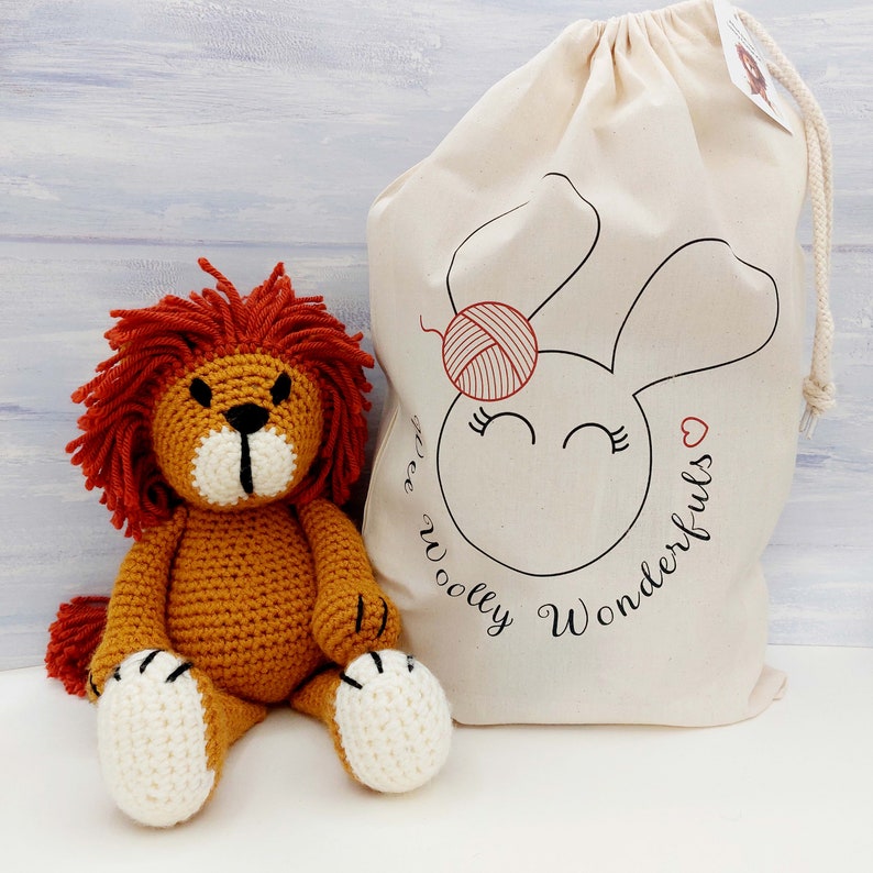 Op de afbeelding: Een gehaakt leeuwen knuffeldier met een rode manen, zittend op een witte canvas tas met een konijnen gezicht en de tekst "Free Woolly Wonderfuls".