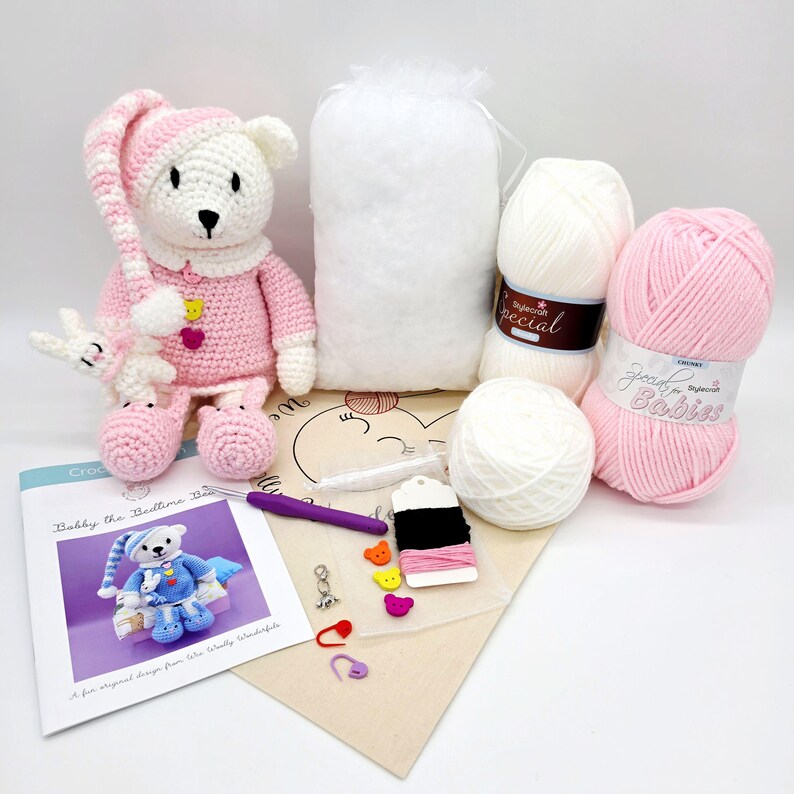 May include: A crochet kit featuring a white and pink stuffed bear, a bag of white stuffing, two skeins of white and pink yarn, a purple crochet hook, small colorful buttons, stitch markers, and an instructional booklet titled Babby the Bedtime Bear.