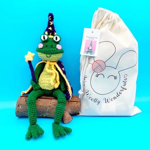 May include: A handmade green crochet frog wearing a wizard hat and cape, holding a star wand, sitting on a log. A drawstring bag with a bunny graphic and the text "Woolly Wonderfuls" is next to the frog. The frog kit includes a tag that says "Fizzy the Wizard Frog Crochet Kit".