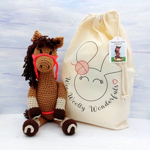 May include: A handmade crochet horse with a brown body, dark brown mane, and a bright pink halter. Beside it is a cream-colored drawstring bag with a bunny graphic and the words "Wee Woolly Wonderfuls". A tag reads "Sundance the Horse Crochet Kit".