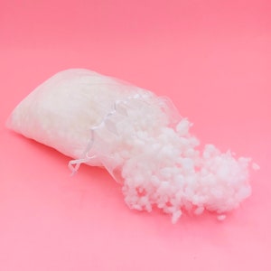 May include: White, fluffy filling spills out of a clear plastic bag onto a pink background. The filling is used for stuffing pillows, cushions, and other soft items.