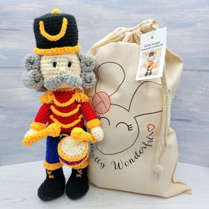 May include: A crocheted Nutcracker doll with a red and gold uniform, holding a yellow and red drum. The doll is packaged in a white canvas bag with a bunny design and the text "Woolly Wonderfuls" and "Ned the Naughty Nutcracker Crochet Kit".