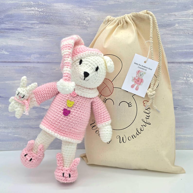 May include: A crocheted white bear doll wearing a pink and white striped nightcap, a pink sweater with three small bear-shaped buttons, and pink slippers. The bear holds a small white crocheted rabbit. A beige drawstring bag with a tag is included.