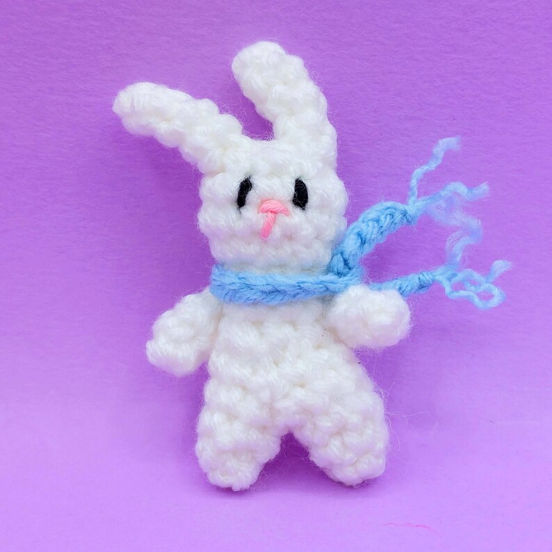 May include: A small white crocheted rabbit figurine featuring two long ears, black embroidered eyes, and a pink stitched nose. The rabbit is wearing a light blue crocheted scarf that extends to the right side with loose yarn ends.