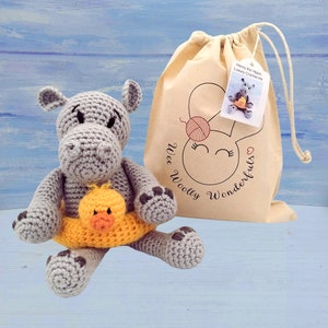 May include: A gray crocheted hippopotamus wearing a yellow crocheted life preserver with a yellow crocheted duck inside. The hippopotamus is sitting on a light blue wooden surface. A white drawstring bag with the text "Wee Woolly Wonderfuls" and "Henry the Hippo Luxury Crochet Kit" is in the background.