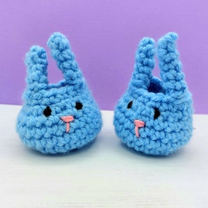 May include: A pair of light blue crochet booties shaped like rabbit heads. Each bootie features two upright ears, small black embroidered eyes, and a tiny pink stitched nose. The textured yarn construction creates a soft, rounded appearance.