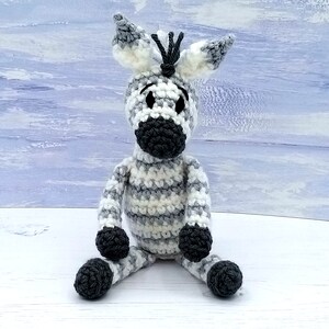 May include: A crocheted zebra toy with black and white stripes, black eyes, and a black nose. The toy is sitting on a white surface.