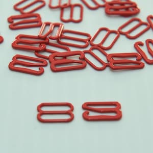 May include: A pile of red metal bra strap adjusters, also known as bra sliders, on a white surface.