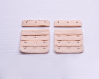 May include: A set of four beige bra extender straps with metal hooks. The straps are designed to extend the length of a bra.