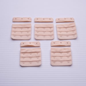 May include: Five beige bra strap extenders with metal hooks and eyes. The extenders are arranged in two rows of three.