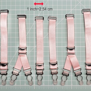 May include: Six pink adjustable garment straps with silver metal clips and clear plastic loops. The straps are laid out on a green grid with a ruler in inches.