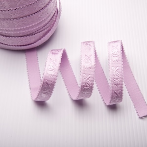 May include: A spool of pale pink satin ribbon with a delicate floral pattern. The ribbon is coiled and a length of ribbon is unfurled and curled on a white surface.