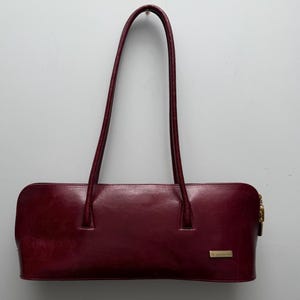 May include: A burgundy leather handbag with two long handles. The rectangular bag has a gold zipper and a small gold-toned plaque. The handles are arched and attached to the top of the bag.