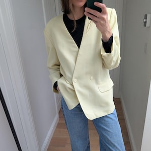 Peut inclure: Un blazer jaune pâle à double boutonnage, porté avec un haut noir et un jean bleu clair à jambes larges. Le blazer a deux boutons et un revers cranté. Des chaussures vernies noires complètent la tenue.