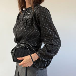May include: A black long-sleeved blouse featuring a white grid pattern, a button-down front, and gathered shoulders, paired with grey trousers and a black woven leather handbag with a thin shoulder strap and a rectangular structured base.