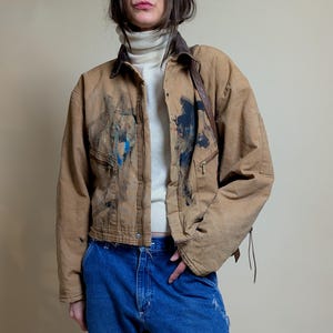 May include: A brown work jacket with paint splatters and a brown collar. The jacket has two zippered pockets and is worn over a white turtleneck. The model is wearing blue jeans.