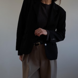 Op de afbeelding: Een zwarte blazer met een klassiek ontwerp, gecombineerd met een donker shirt en een beige broek. De blazer heeft een revers en een enkele knoopsluiting. Een bruine riem met een gouden gesp is zichtbaar. De outfit is eenvoudig en elegant.