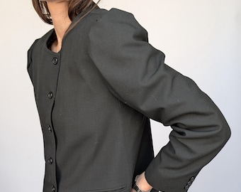 May include: A black blazer with a round collar and button closure. The blazer has a single pocket and long sleeves. The sleeves have buttons at the cuffs. The blazer is paired with black pants.