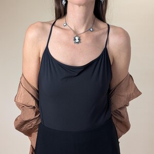 May include: A woman wearing a black camisole and black pants. She is wearing a silver necklace with a cameo pendant and matching earrings. A brown jacket is draped over her shoulders. The background is a solid beige color.