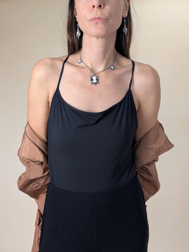 May include: A woman wearing a black camisole and black pants. She is wearing a silver necklace with a cameo pendant and matching earrings. A brown jacket is draped over her shoulders. The background is a solid beige color.