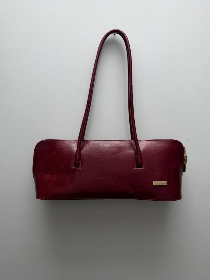 May include: A burgundy leather handbag with two long handles. The rectangular bag has a gold zipper and a small gold-toned plaque. The handles are arched and attached to the top of the bag.