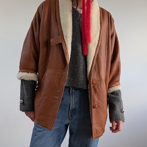 May include: A brown leather jacket with a cream-colored shearling collar and cuffs. The jacket has a button closure and a pocket on the left side. The sleeves are gray knit with silver buttons. The person is wearing blue jeans.