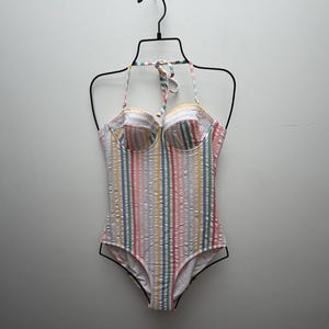 May include: A one-piece swimsuit featuring a textured seersucker fabric with vertical stripes in shades of pink, yellow, blue, and green. The garment includes a sweetheart neckline, molded cups, and thin halter-style straps that tie at the neck.