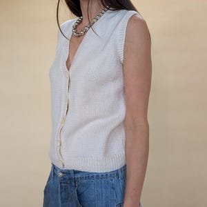 Peut inclure: Un pull sans manches en tricot blanc cass&eacute; avec un col en V et une fermeture boutonn&eacute;e. Le pull est associ&eacute; &agrave; un jean en denim bleu. Un collier de perles argent&eacute;es est port&eacute; autour du cou. L'ensemble est d&eacute;contract&eacute;.