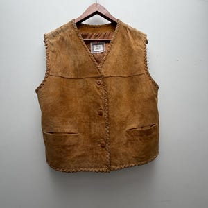 May include: A tan suede vest with a V-neck, three buttons, and two front pockets. The edges are trimmed with a decorative stitch. The vest is hanging on a wooden hanger.