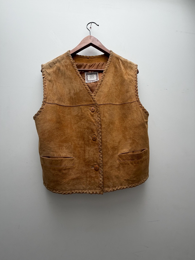 May include: A tan suede vest with a V-neck, three buttons, and two front pockets. The edges are trimmed with a decorative stitch. The vest is hanging on a wooden hanger.