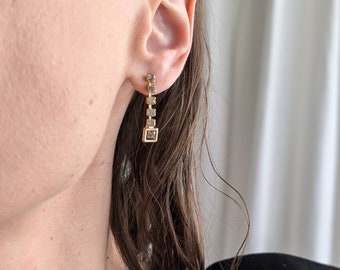 May include: A pair of gold earrings with a dangling chain of small square-shaped stones. The stones are a light brown color.