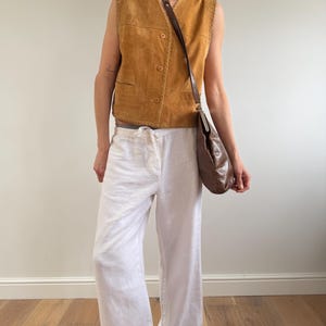 May include: A tan suede vest with a button-up front and a brown leather shoulder bag. The outfit includes white linen pants. The vest has decorative stitching along the edges. The bag has a long strap.