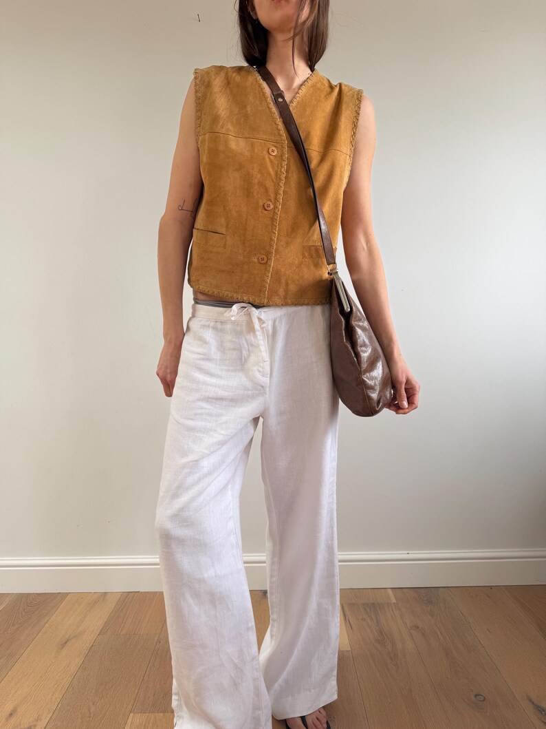 May include: A tan suede vest with a button-up front and a brown leather shoulder bag. The outfit includes white linen pants. The vest has decorative stitching along the edges. The bag has a long strap.