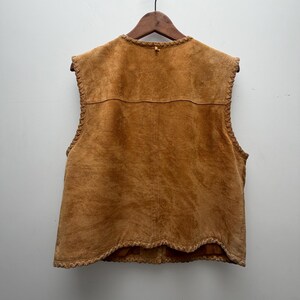 May include: A tan suede vest with a round neckline and a decorative trim along the edges. The vest is sleeveless and hangs on a wooden hanger against a white background. The back of the vest is visible, showing the stitching and texture of the suede.