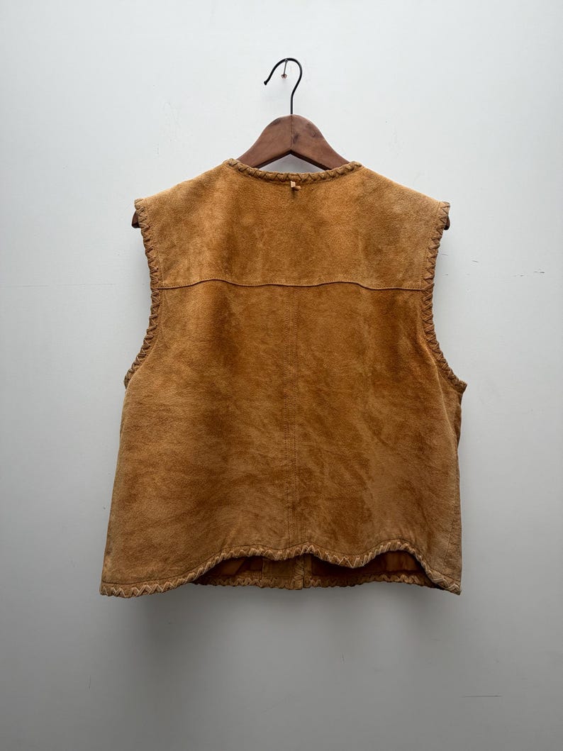 May include: A tan suede vest with a round neckline and a decorative trim along the edges. The vest is sleeveless and hangs on a wooden hanger against a white background. The back of the vest is visible, showing the stitching and texture of the suede.