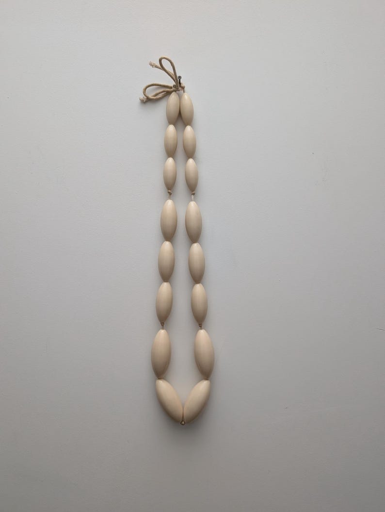 May include: Cream-colored beaded necklace with oval-shaped beads. The beads are strung together with a natural-colored cord, forming a long, elegant design. The necklace is displayed against a plain white background.