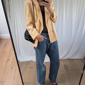 Op de afbeelding: Een lichtperzik blazer met gouden knopen, gecombineerd met een blauwe jeans en zwarte pumps met spitse neus. Een zwarte handtas hangt over de schouder. De outfit wordt weerspiegeld in een spiegel.