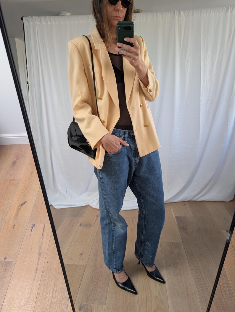 Op de afbeelding: Een lichtperzik blazer met gouden knopen, gecombineerd met een blauwe jeans en zwarte pumps met spitse neus. Een zwarte handtas hangt over de schouder. De outfit wordt weerspiegeld in een spiegel.