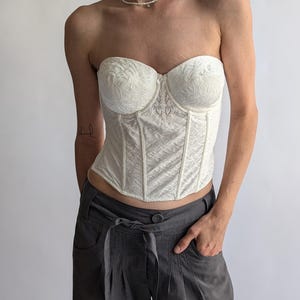May include: White strapless corset with a floral lace design. The corset has a boned structure and a hook and eye closure in the back.