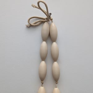 May include: A strand of six off-white, oval-shaped beads, connected in pairs by thin, beige string. The top of the strand is tied with a loop of beige string and secured with a small metal pin. The background is a plain white surface.