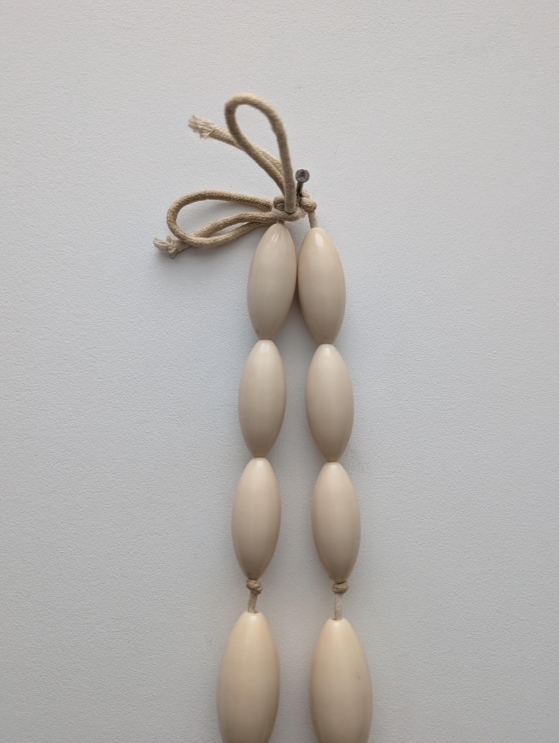 May include: A strand of six off-white, oval-shaped beads, connected in pairs by thin, beige string. The top of the strand is tied with a loop of beige string and secured with a small metal pin. The background is a plain white surface.