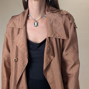 May include: A light brown trench coat with large lapels and two visible buttons. A black top is worn underneath. A silver necklace and earrings with cameo pendants are also visible.